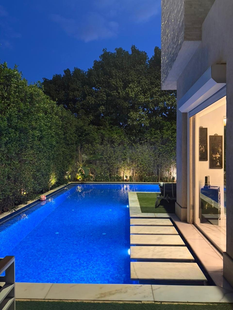 Lit swimming pool at dusk with marble stepping stones beside the house