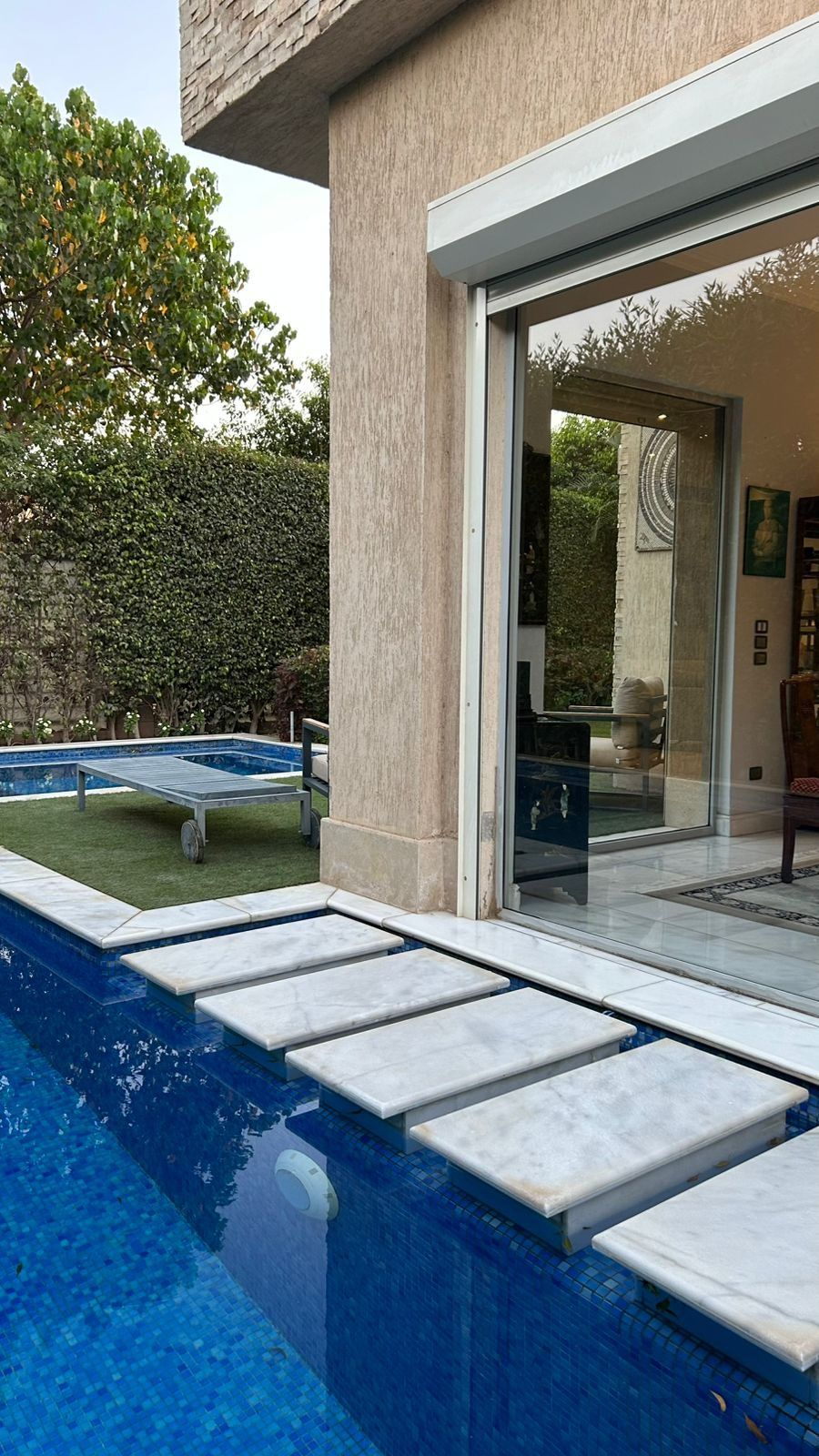 Marble stepping stones over a blue mosaic pool next to floor-to-ceiling glazing